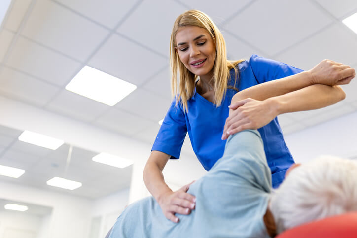 Female physical therapist stretching the arm and shoulder of a senior woman on a treatment table. Professional rehabilitation session for elderly mobility in a bright modern medical clinic.